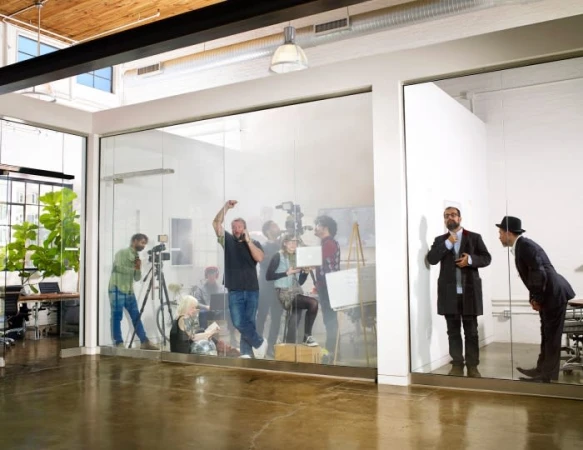 Behind glass windows, a video and photo shoot in progress featuring a diverse group of adults, with two businessmen observing.