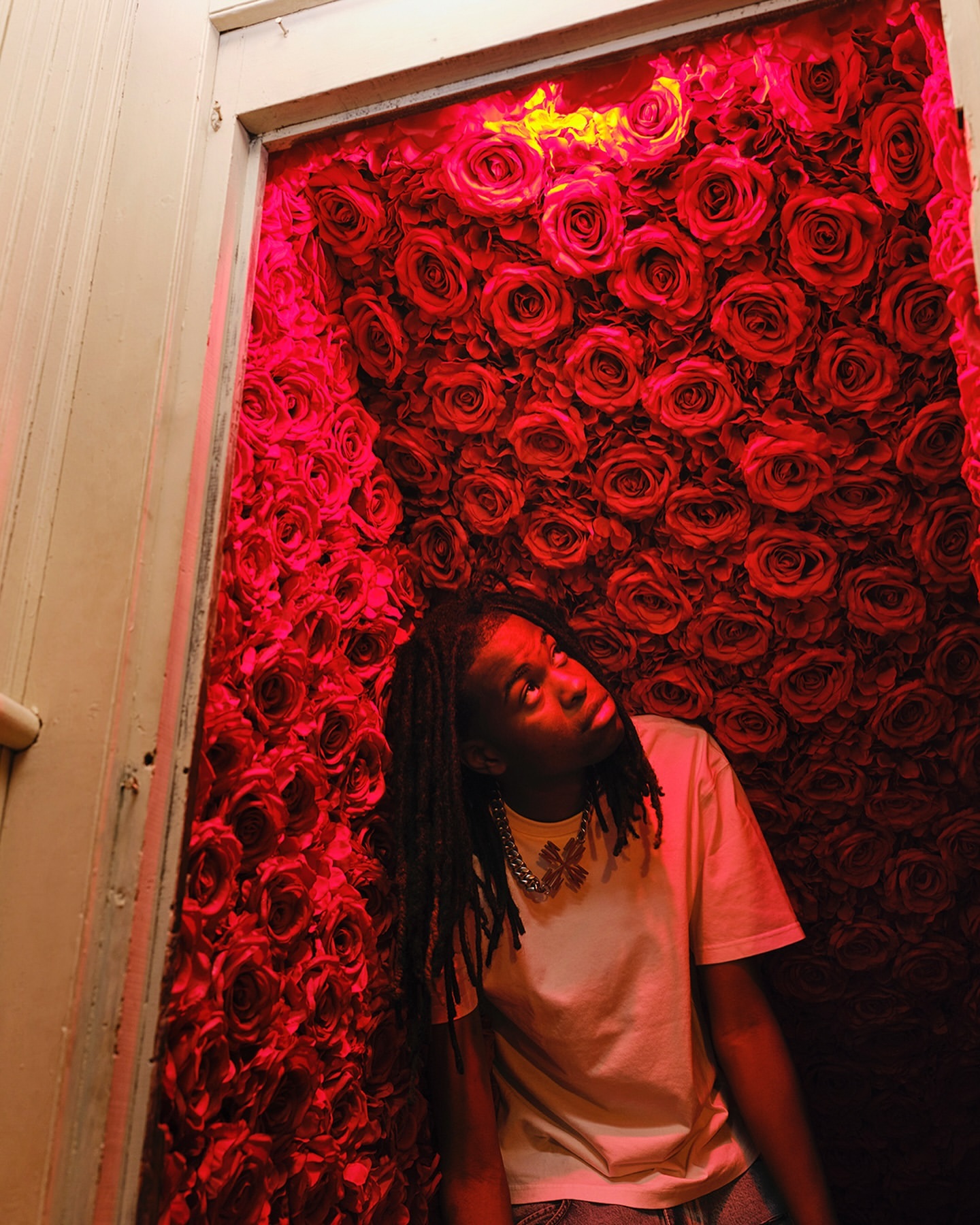 Young adult poses in front of a vibrant wall of red roses, bathed in a red light, wearing a white t-shirt and silver butterfly necklace.