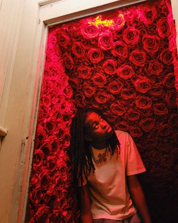Young adult poses in front of a vibrant wall of red roses, bathed in a red light, wearing a white t-shirt and silver butterfly necklace.