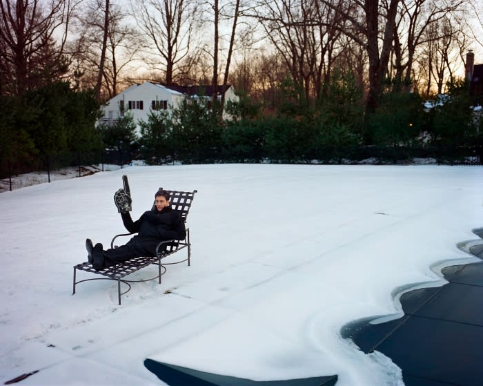 Adult male in winter wear lounges on a metal chair in a snow-covered yard, holding a foam finger with houses and trees in the background.