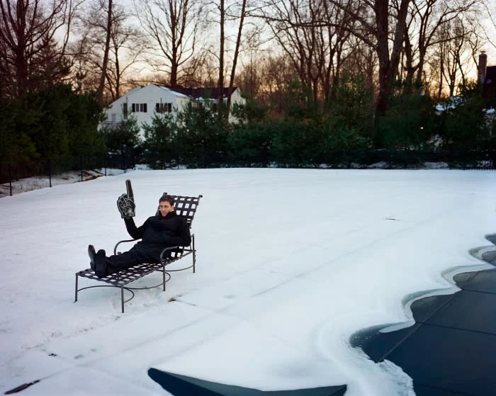 Adult male in winter wear lounges on a metal chair in a snow-covered yard, holding a foam finger with houses and trees in the background.