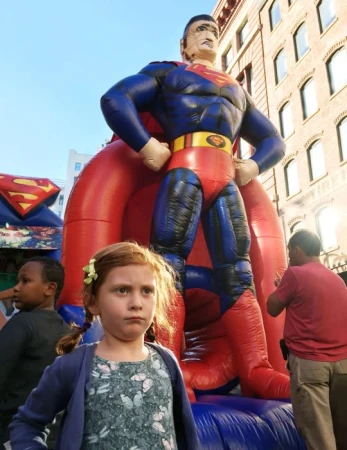 A young red-haired girl stands in front of an inflatable Superman, with buildings and sky in the background, at an outdoor event.