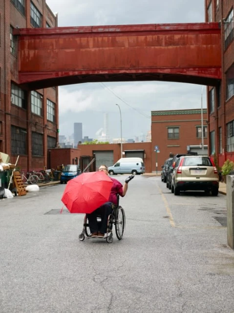Adult in wheelchair holding a red umbrella on a city street between red brick warehouse buildings with metal industrial skyway connector.