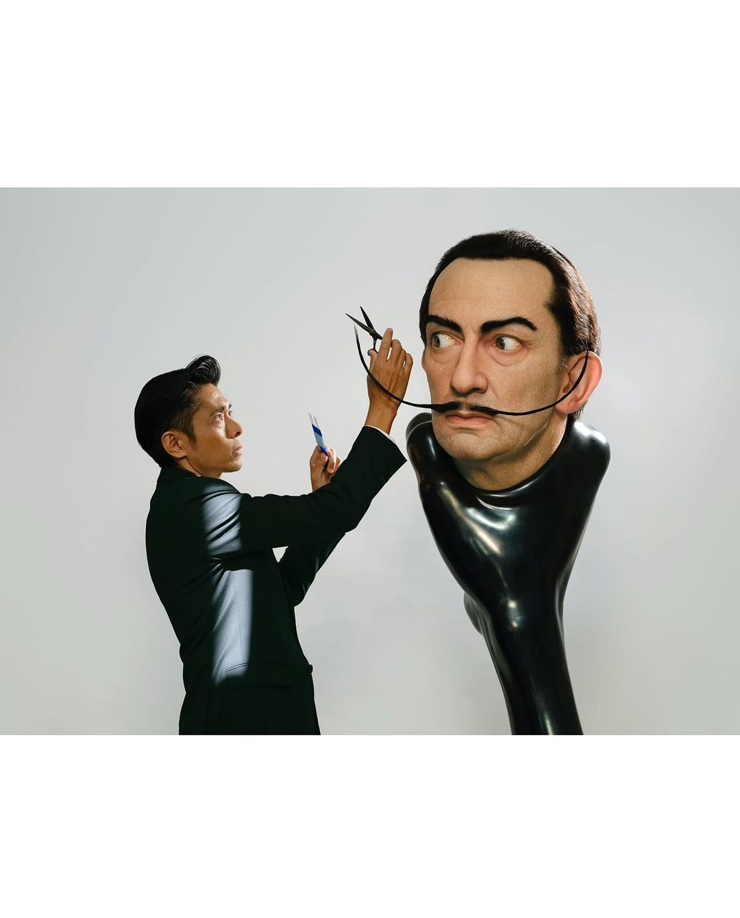 Asian adult male cuts the mustache of a mannequin of a male's head with scissors in a photo studio with a white background.
