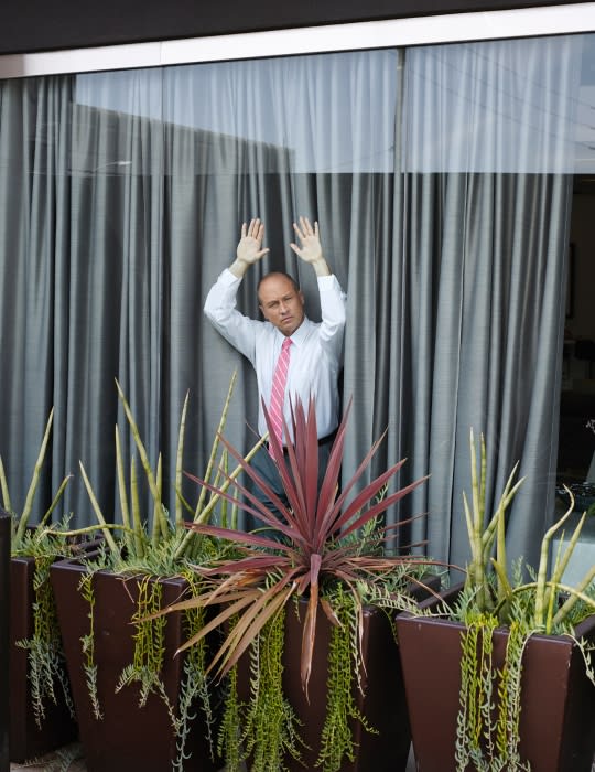 Adult male with hands up in front of a window, framed by potted plants and grey curtains, wearing a white shirt and pink tie.
