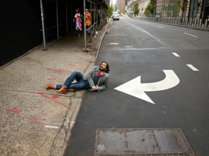 Adult male lying on city street next to white painted arrow, wearing jeans and a jacket. Buildings are in the background.