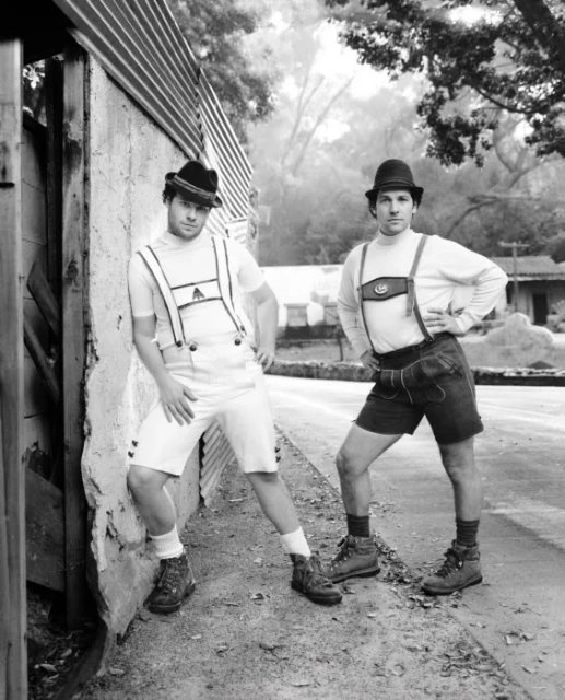 Two adult males in traditional Bavarian outfits pose outdoors on a paved path with a building on the left and trees in the background.