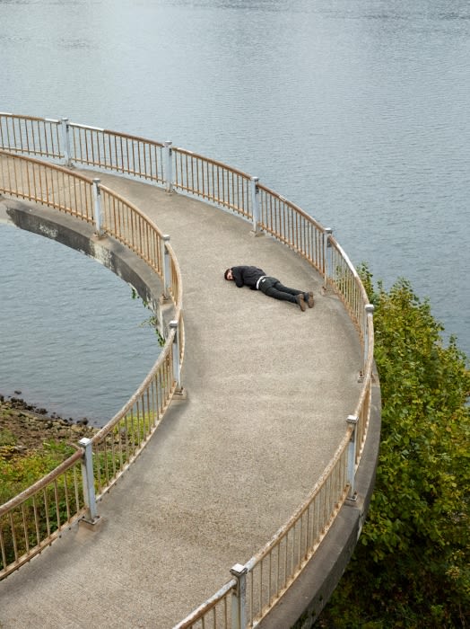Person lies prone on a curved concrete walkway overlooking a body of water with green trees visible to the lower right.
