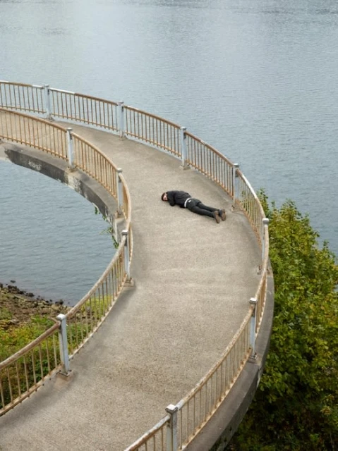 Person lies prone on a curved concrete walkway overlooking a body of water with green trees visible to the lower right.