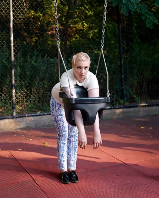 Adult female stands beside a black child's swing in a playground with a chain-link fence and trees visible in the background on a sunny day.