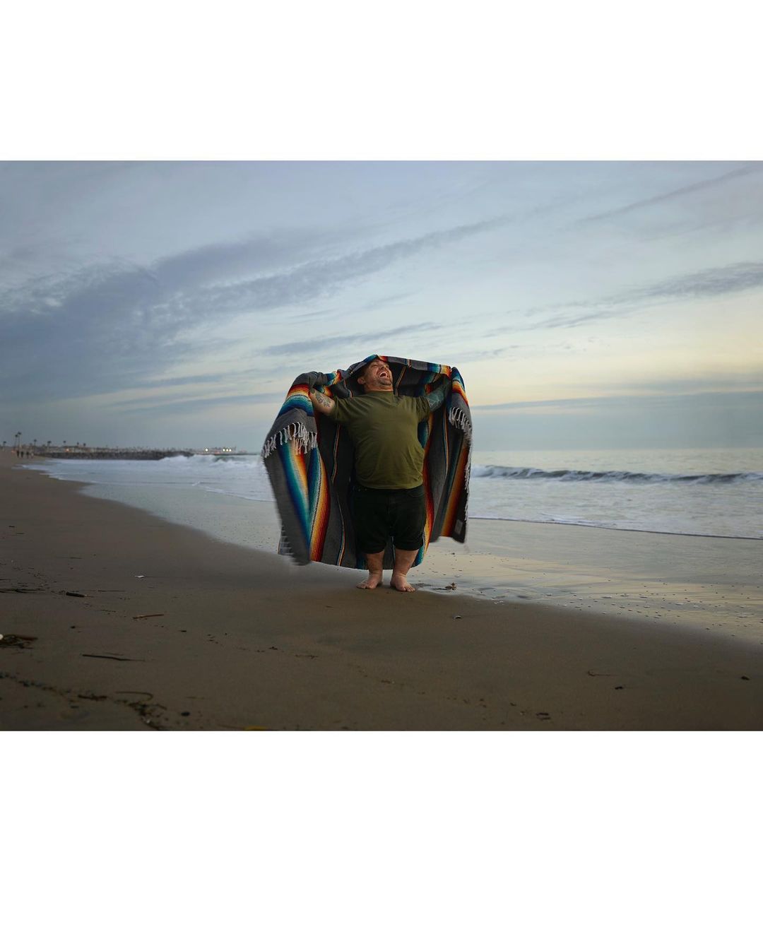 Person wrapped in a colorful blanket stands on a sandy beach, with the ocean and cloudy sky in the background.