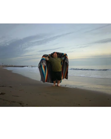 Person wrapped in a colorful blanket stands on a sandy beach, with the ocean and cloudy sky in the background.