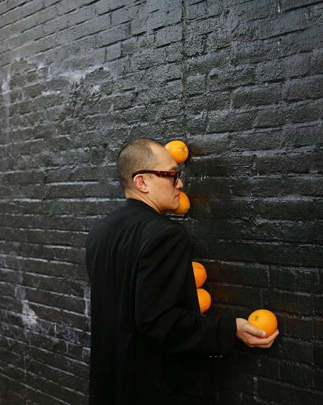 Adult male in a black suit places oranges against a black brick wall in an artistic display. The composition is simple and striking.