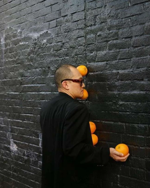 Adult male in a black suit places oranges against a black brick wall in an artistic display. The composition is simple and striking.