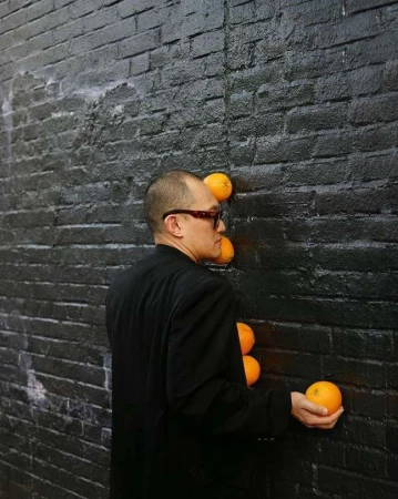Adult male in a black suit places oranges against a black brick wall in an artistic display. The composition is simple and striking.