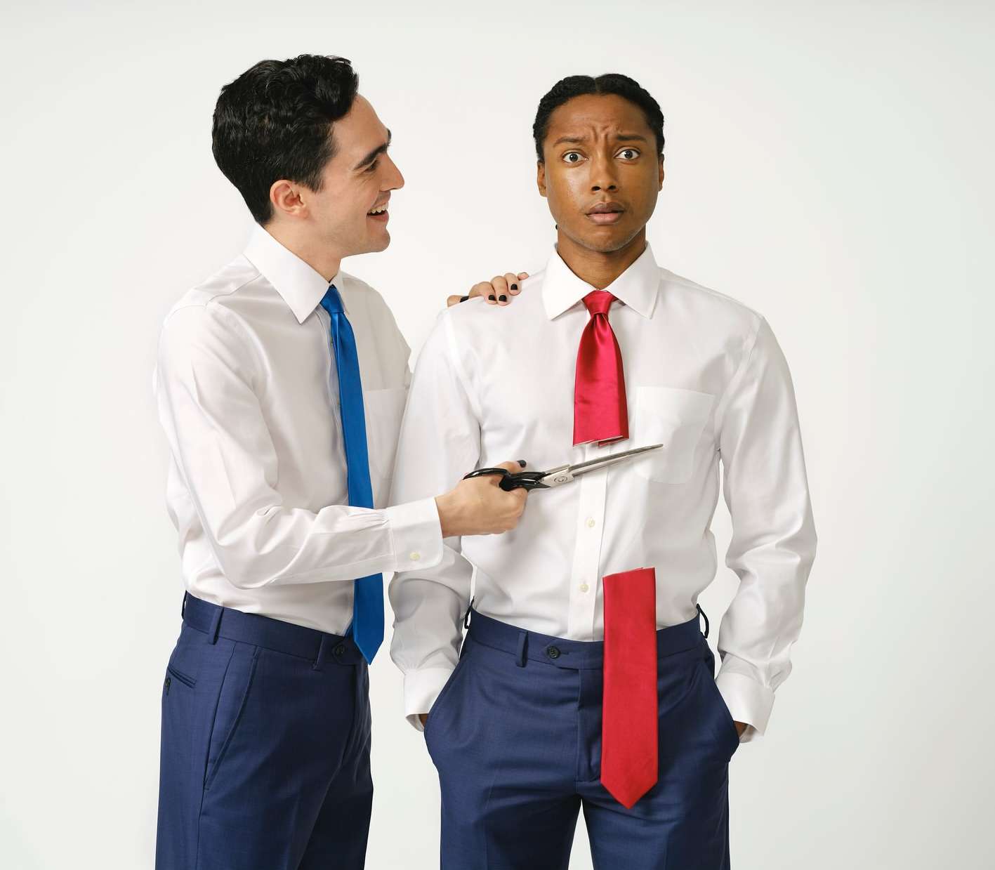 Two men in a bright photo studio, one man cutting the other man's necktie with scissors; both wear white dress shirts and blue trousers.