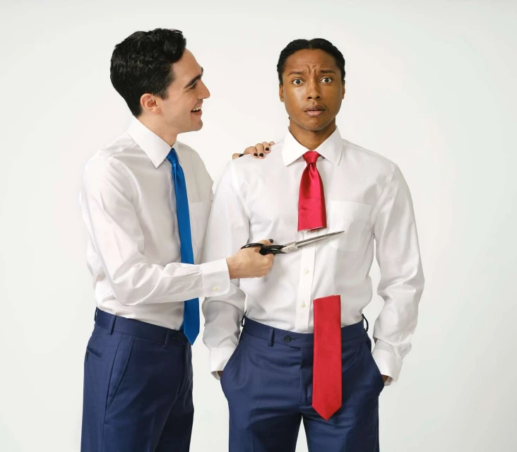Two men in a bright photo studio, one man cutting the other man's necktie with scissors; both wear white dress shirts and blue trousers.