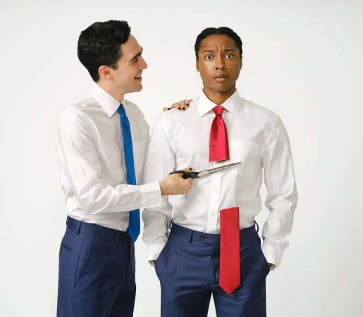Two men in a bright photo studio, one man cutting the other man's necktie with scissors; both wear white dress shirts and blue trousers.