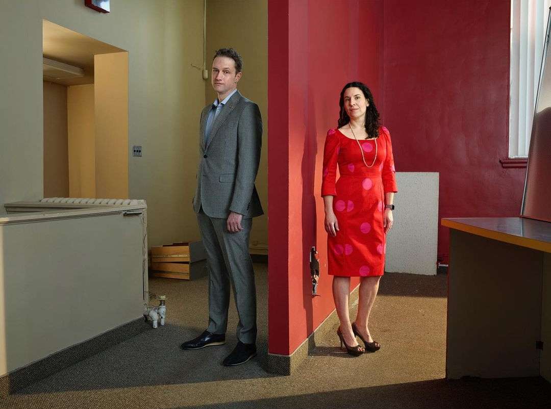 Indoor portrait of a man in a suit and a woman in a polka-dot dress standing on either side of a red wall, looking directly at the camera.