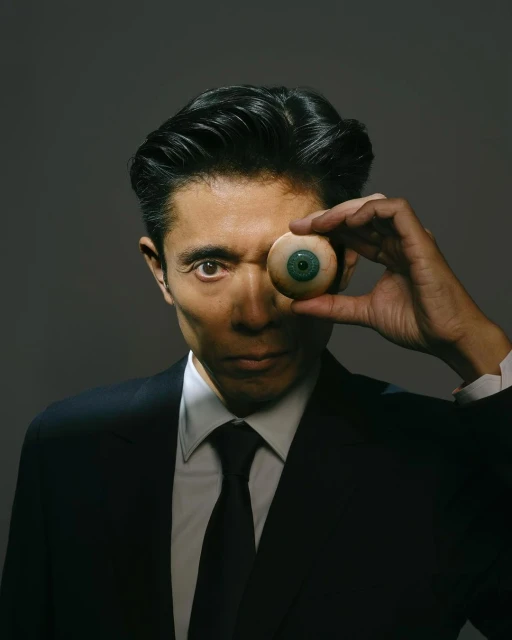 Person in suit poses with a prosthetic eye in hand against a grey background. The adult male looks directly at the viewer.