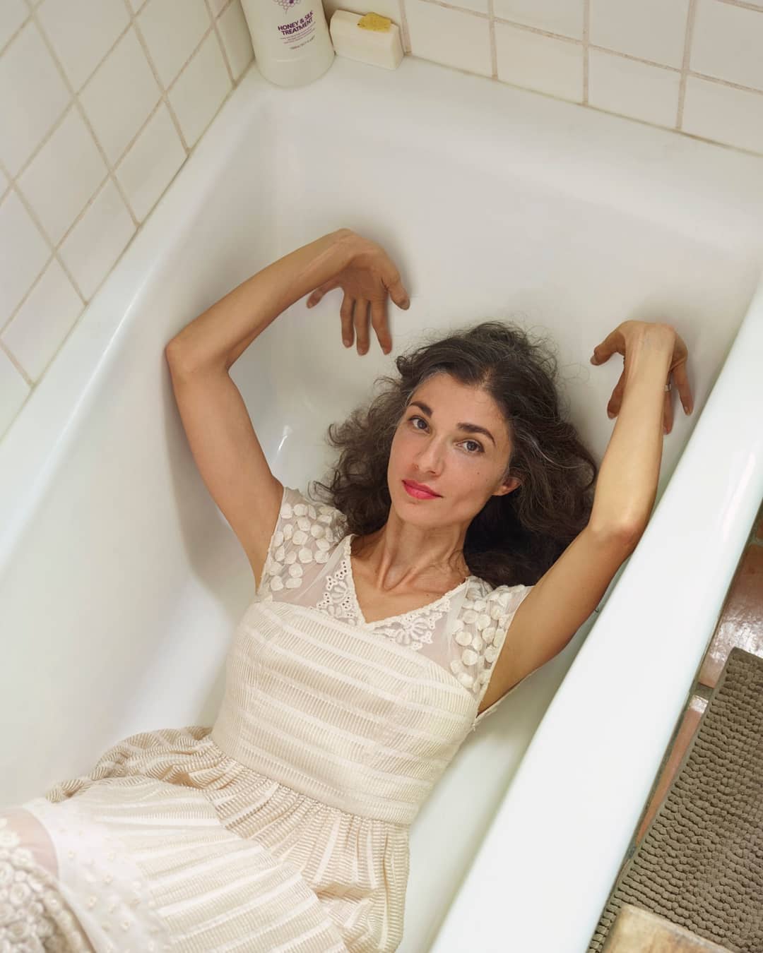 Woman in a light-colored dress poses in a bathtub, looking directly at the camera with bright red lipstick and neutral expression.