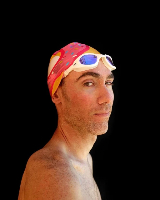 Profile of a man wearing a donut-patterned swim cap and swimming goggles against a stark black background. Studio lighting highlights the details.