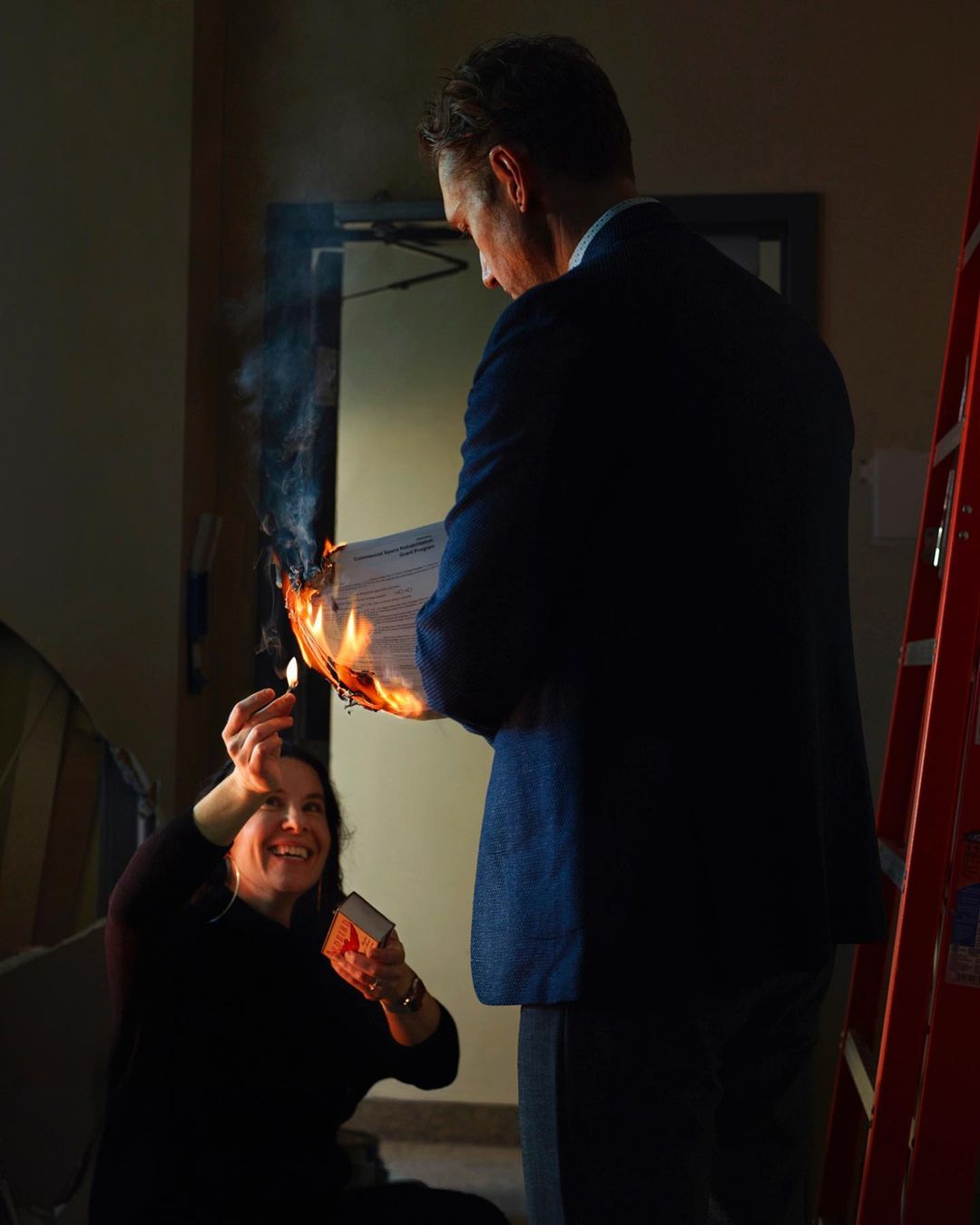 Two people enacting a scene indoors, where an adult male holds a flaming document, and an adult female holds a box of matches.