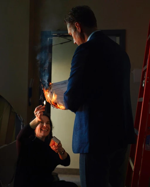 Two people enacting a scene indoors, where an adult male holds a flaming document, and an adult female holds a box of matches.