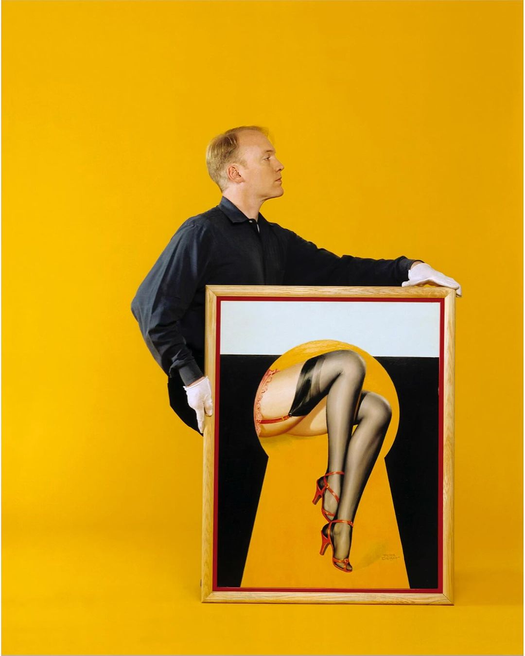 Man in photo studio holding framed artwork of woman's legs in black stockings and red high heels against a yellow background.