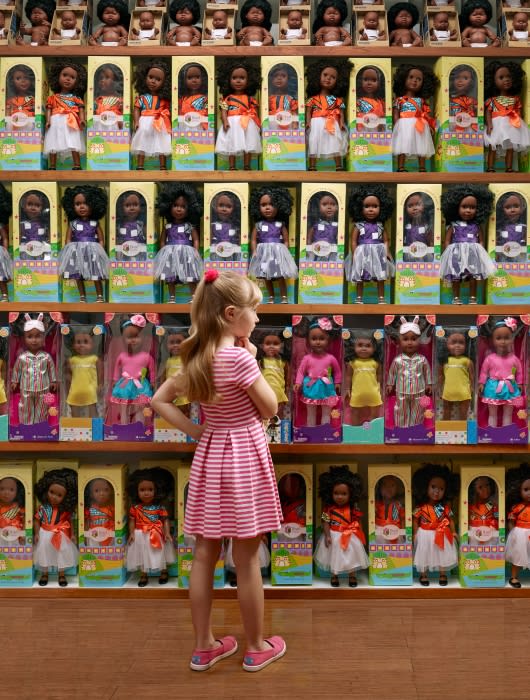Young girl in pink stands in front of a store shelf filled with African American fashion dolls in boxes in a brightly lit store.