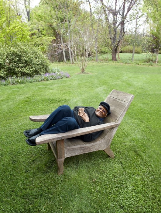 An African-American man wearing a black hat and shirt, reclines in an adirondack chair on a green lawn, surrounded by trees in daylight.
