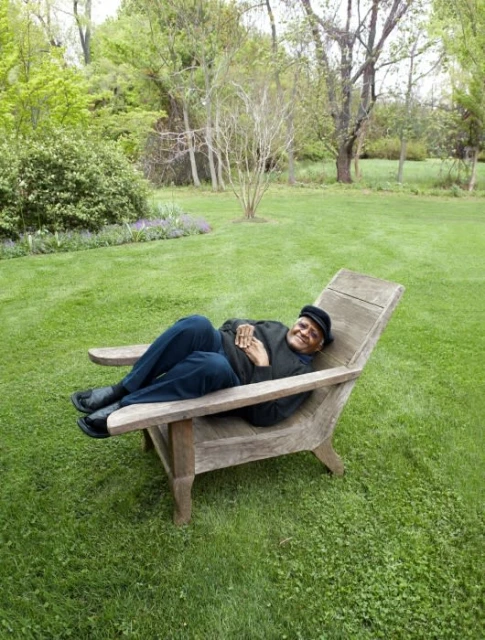 An African-American man wearing a black hat and shirt, reclines in an adirondack chair on a green lawn, surrounded by trees in daylight.