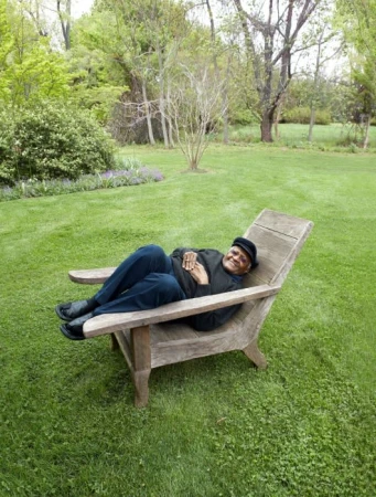 An African-American man wearing a black hat and shirt, reclines in an adirondack chair on a green lawn, surrounded by trees in daylight.