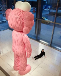 Giant pink art sculpture indoors facing a person seated on tiled floor near glass windows overlooking a city street