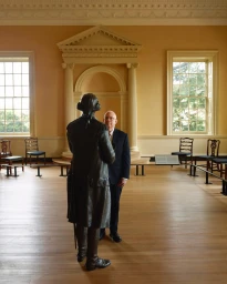 Man in suit standing behind a bronze historical statue inside a large wood-floored hall with yellow walls.