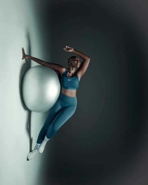 Athletic bald Black woman in sportswear poses with arms outstretched against large silver exercise ball in a fitness fashion shot.
