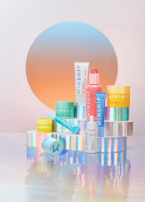 Assortment of Kinship brand skincare products including cleanser, serum, and moisturizer arranged on iridescent platforms against gradient backdrop.