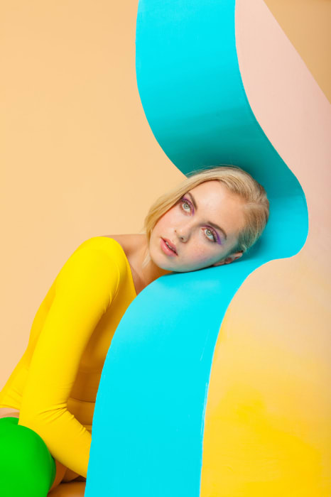 Blonde woman in yellow bodysuit poses by turquoise and peach arches in a brightly lit indoor studio setting for a fashion-forward photo.