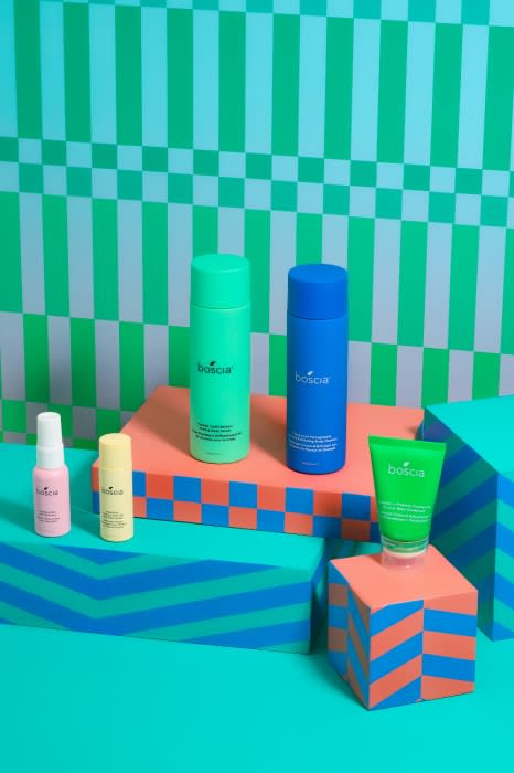 Studio shot of Boscia skincare products, including a spray bottle, a balm stick, a green tube, and two pump bottles, on geometric platforms.