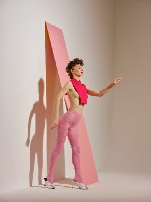 A woman in pink patterned leggings and a bright pink top poses next to a pink triangular geometric shape in a light neutral studio.
