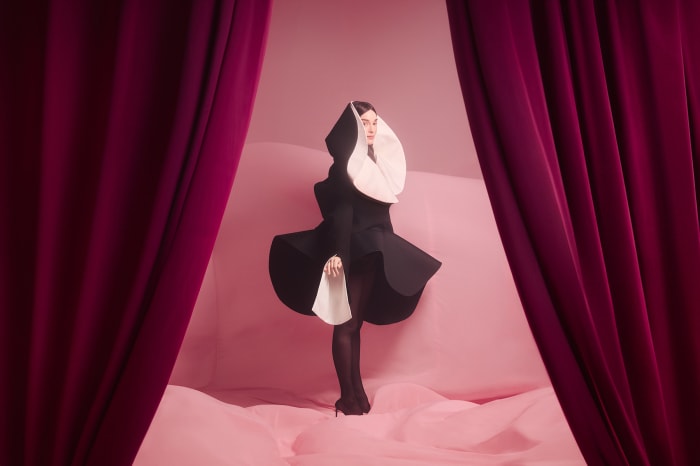 Woman in black and white stylized nun's habit standing indoors on pink backdrop framed by burgundy curtains, dark colors, studio lighting.
