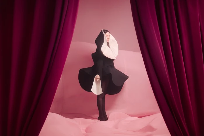 Woman in black and white stylized nun's habit standing indoors on pink backdrop framed by burgundy curtains, dark colors, studio lighting.