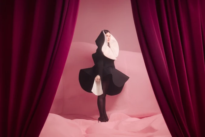 Woman in black and white stylized nun's habit standing indoors on pink backdrop framed by burgundy curtains, dark colors, studio lighting.
