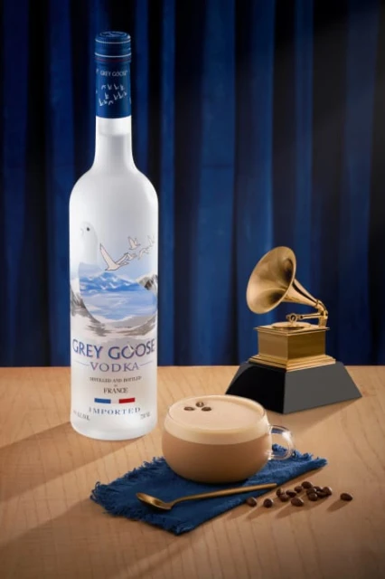 Grey Goose vodka bottle, coffee cocktail, gramophone, and coffee beans on a wood table with a blue curtain backdrop. Still life.