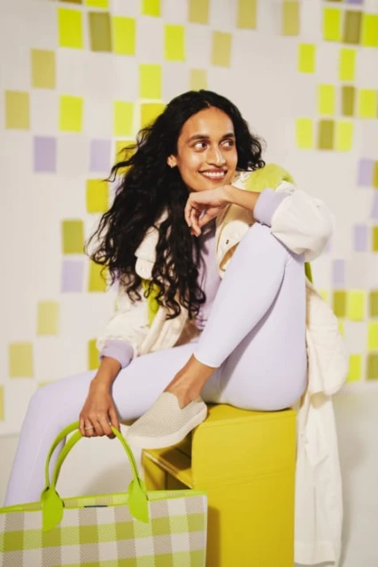 A brunette woman poses in lilac leggings and an off-white jacket, sitting on a yellow block holding a green plaid tote bag.