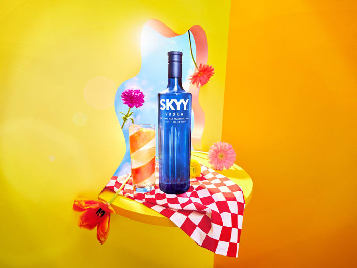 Still life of Skyy Vodka bottle, cocktail, and flowers on a platform against a vibrant yellow and orange background.