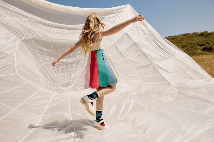 Young girl in colorful dress leaps in front of large white fabric against an outdoor background with blue sky and green hills.