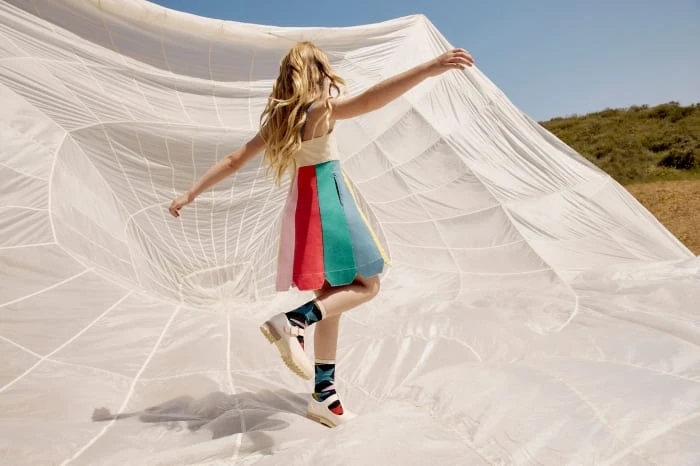 Young girl in colorful dress leaps in front of large white fabric against an outdoor background with blue sky and green hills.