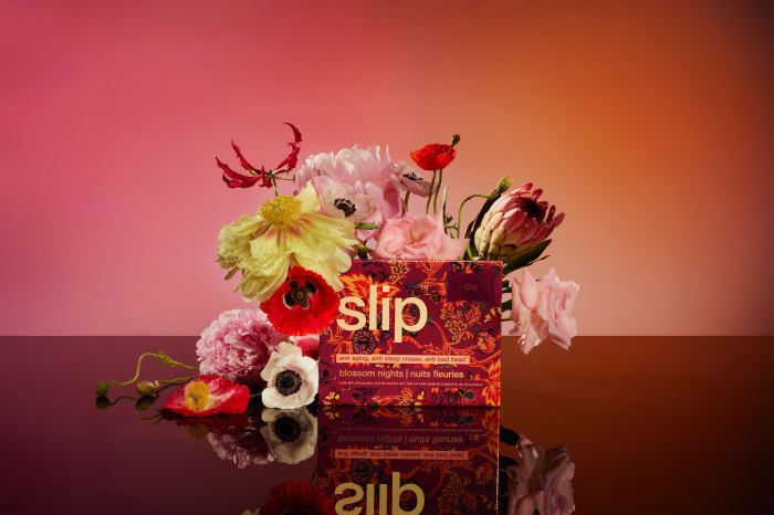 Slip Blossom Nights product box surrounded by colorful flowers on a reflective surface and warm gradient background.