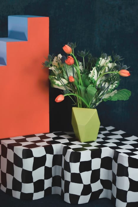 Still life featuring a green vase with orange tulips, and white floral accents set on a black and white checkered surface.